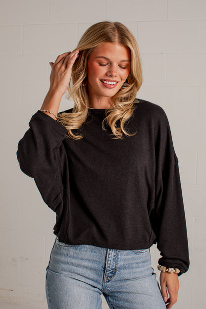 A woman wearing a black long-sleeve crewneck sweatshirt and light wash blue jeans, standing against a white brick wall.