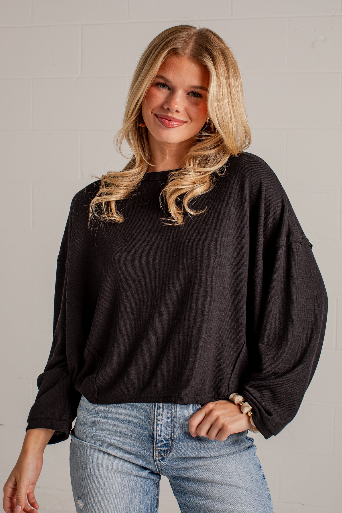 A woman wearing a black oversized long-sleeve sweatshirt and light blue jeans, standing against a light gray brick wall.