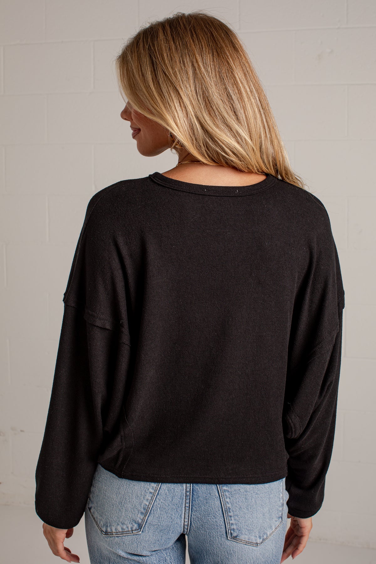 A woman viewed from behind wearing a black long-sleeve crewneck top and light blue jeans, standing against a light gray brick wall.