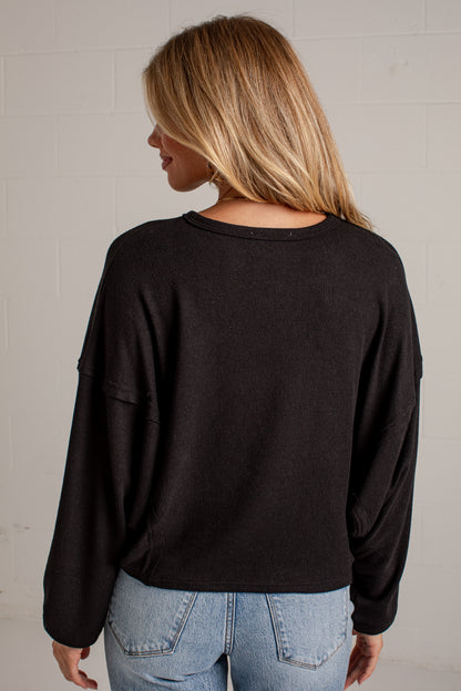 A woman viewed from behind wearing a black long-sleeve crewneck top and light blue jeans, standing against a light gray brick wall.