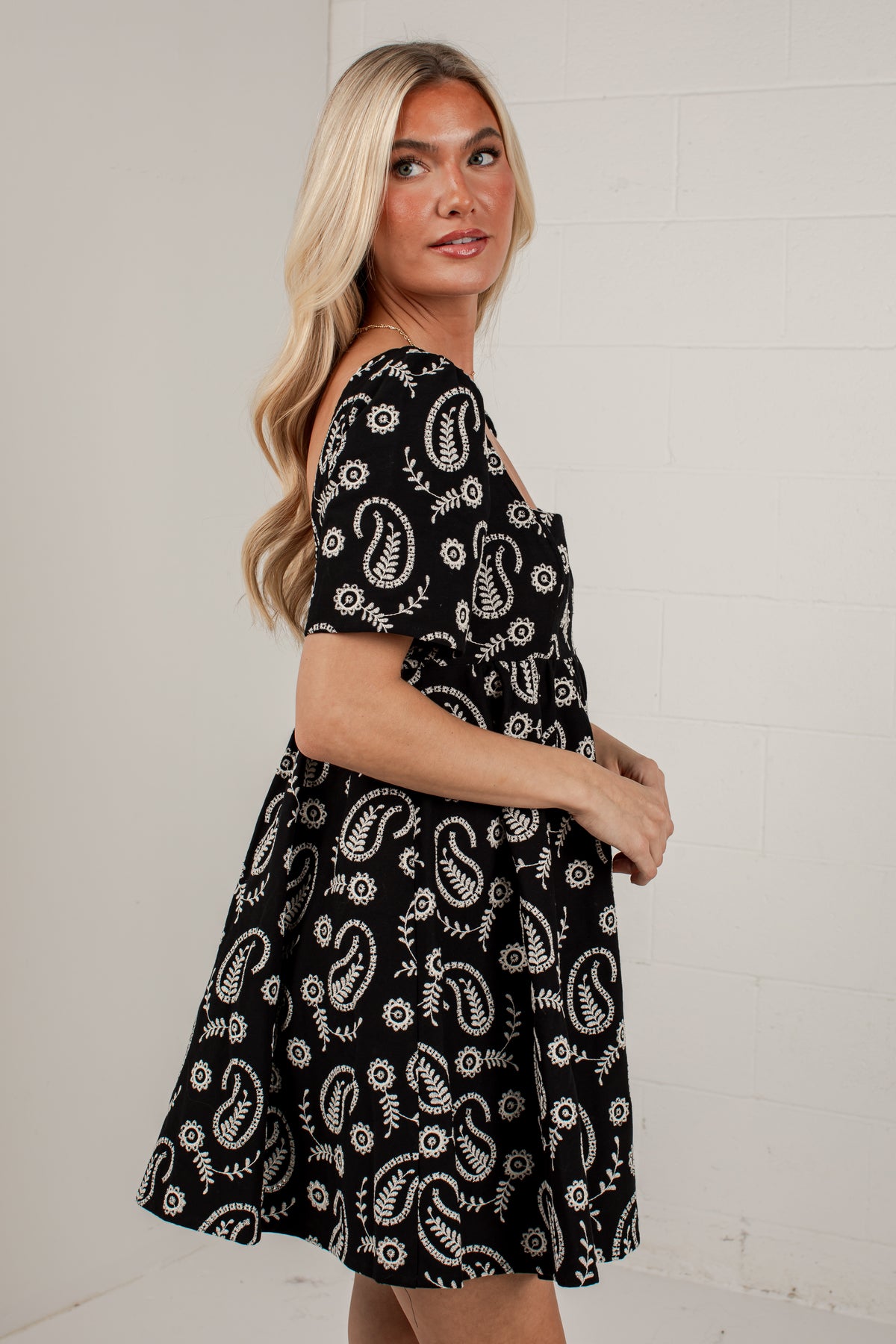 A woman modeling a short-sleeve black dress with a white paisley and floral block print, standing against a white brick wall.