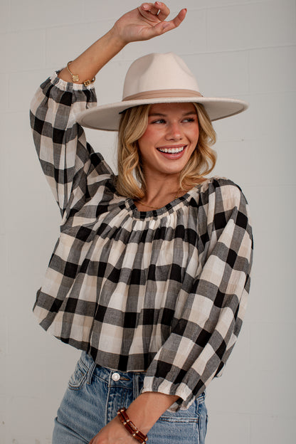 A woman wearing a black and beige buffalo plaid long-sleeve blouse with a gathered neckline, paired with a beige wide-brim hat and blue jeans, posing against a white paneled wall.