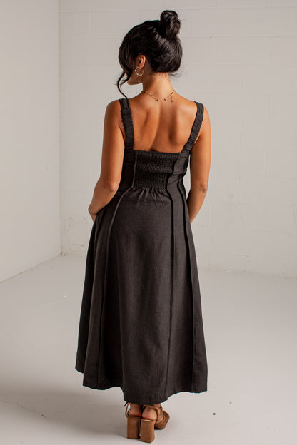 A woman viewed from behind wearing a dark charcoal gray midi dress with shoulder straps, a smocked bodice, and a flared skirt, paired with brown block-heel sandals in a bright studio setting.