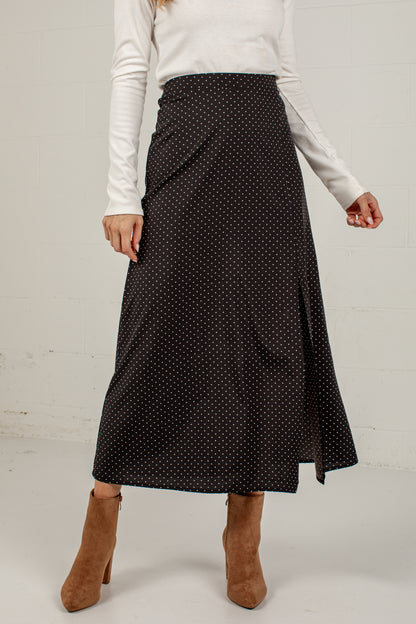 A woman wearing a black midi skirt with a small white polka dot pattern, styled with a white long-sleeve top and brown suede ankle boots against a white brick wall.