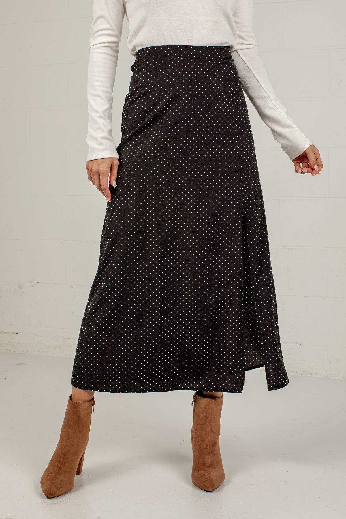 A woman wearing a black midi skirt with a small white polka dot pattern, styled with a white long-sleeve top and brown suede ankle boots against a white studio background.