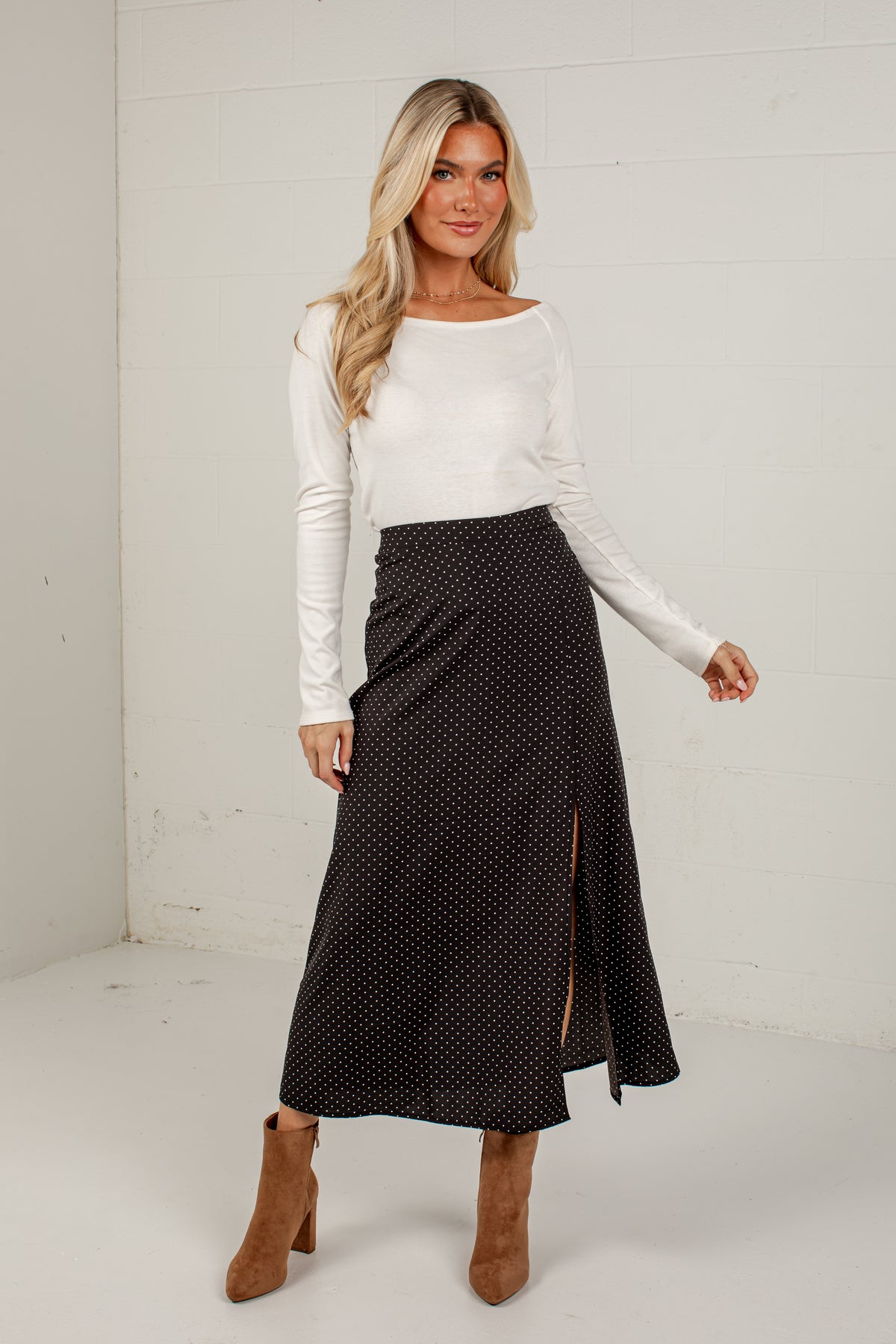 A woman modeling a black midi skirt with small white polka dots and a side slit, paired with a white long-sleeve top and brown ankle boots, standing against a white paneled wall.