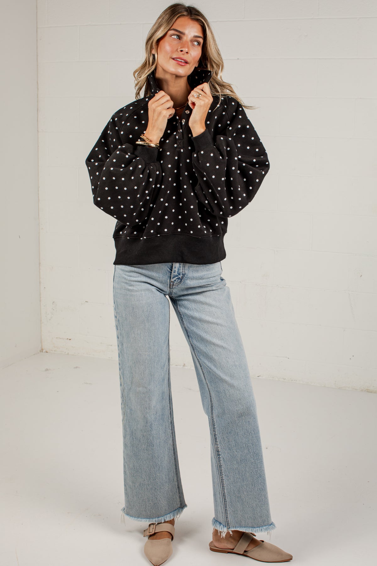 A woman modeling a black long-sleeve top with white polka dots, paired with light-wash wide-leg jeans and beige flats, standing against a white wall.