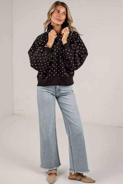A woman modeling a black long-sleeve top with white polka dots, paired with light-wash wide-leg jeans and beige flats, standing against a white wall.