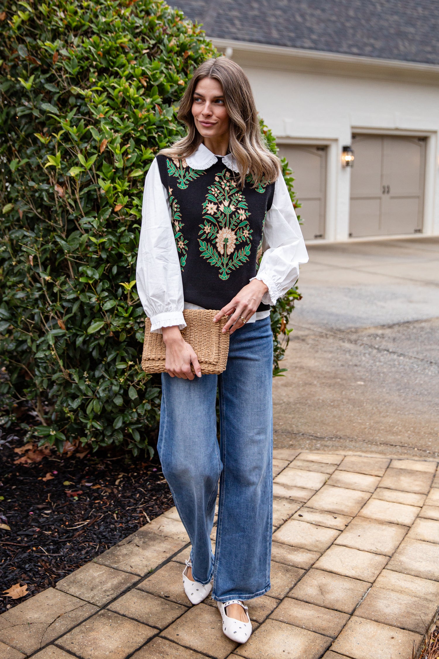 Woman wearing a black embroidered vest over a white blouse with blue jeans, standing on a driveway.