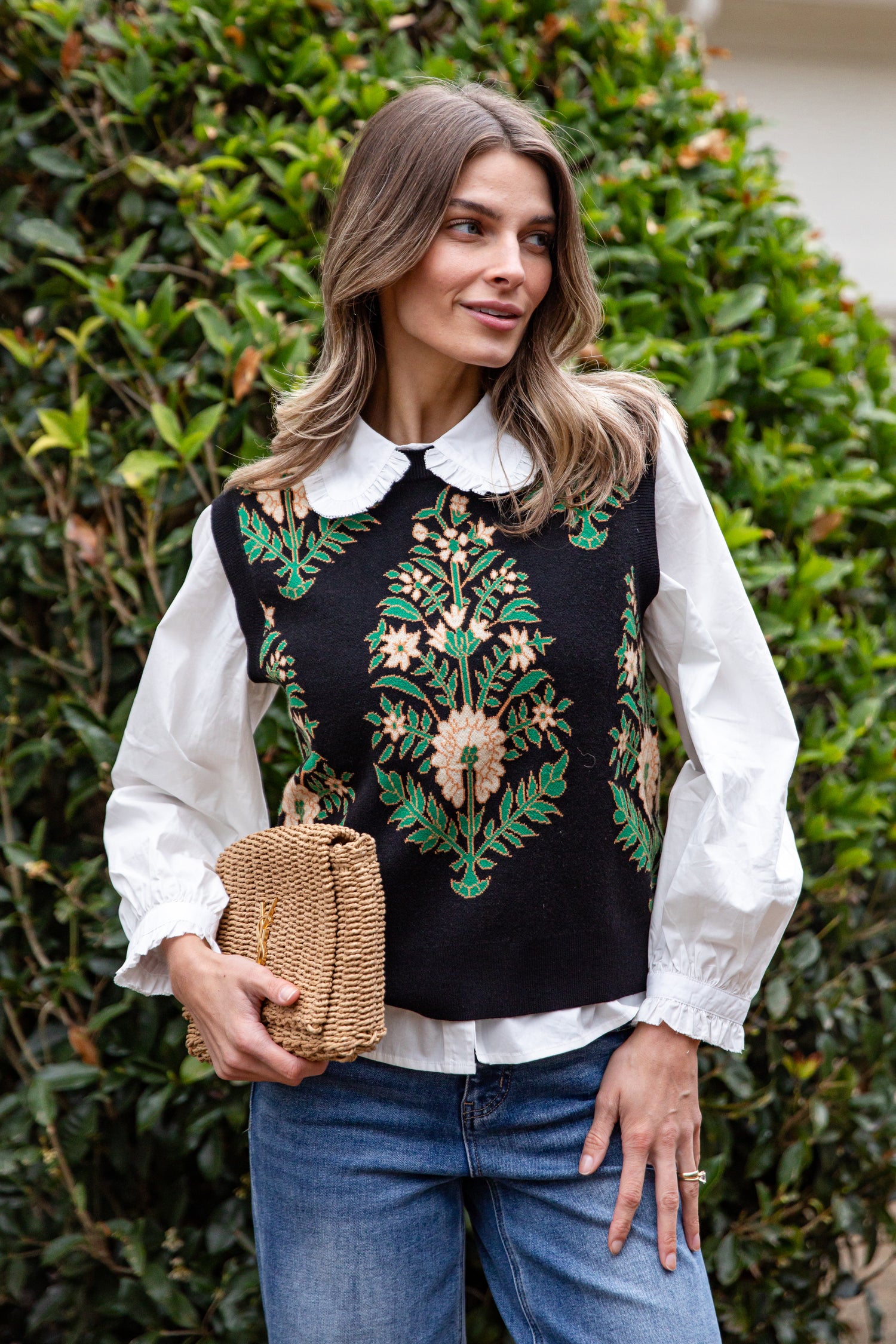 Woman wearing a black embroidered vest over a white blouse with blue jeans, standing against a green leafy background.