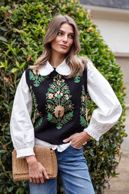 Woman wearing a black embroidered vest over a white blouse with blue jeans, standing in front of green foliage.