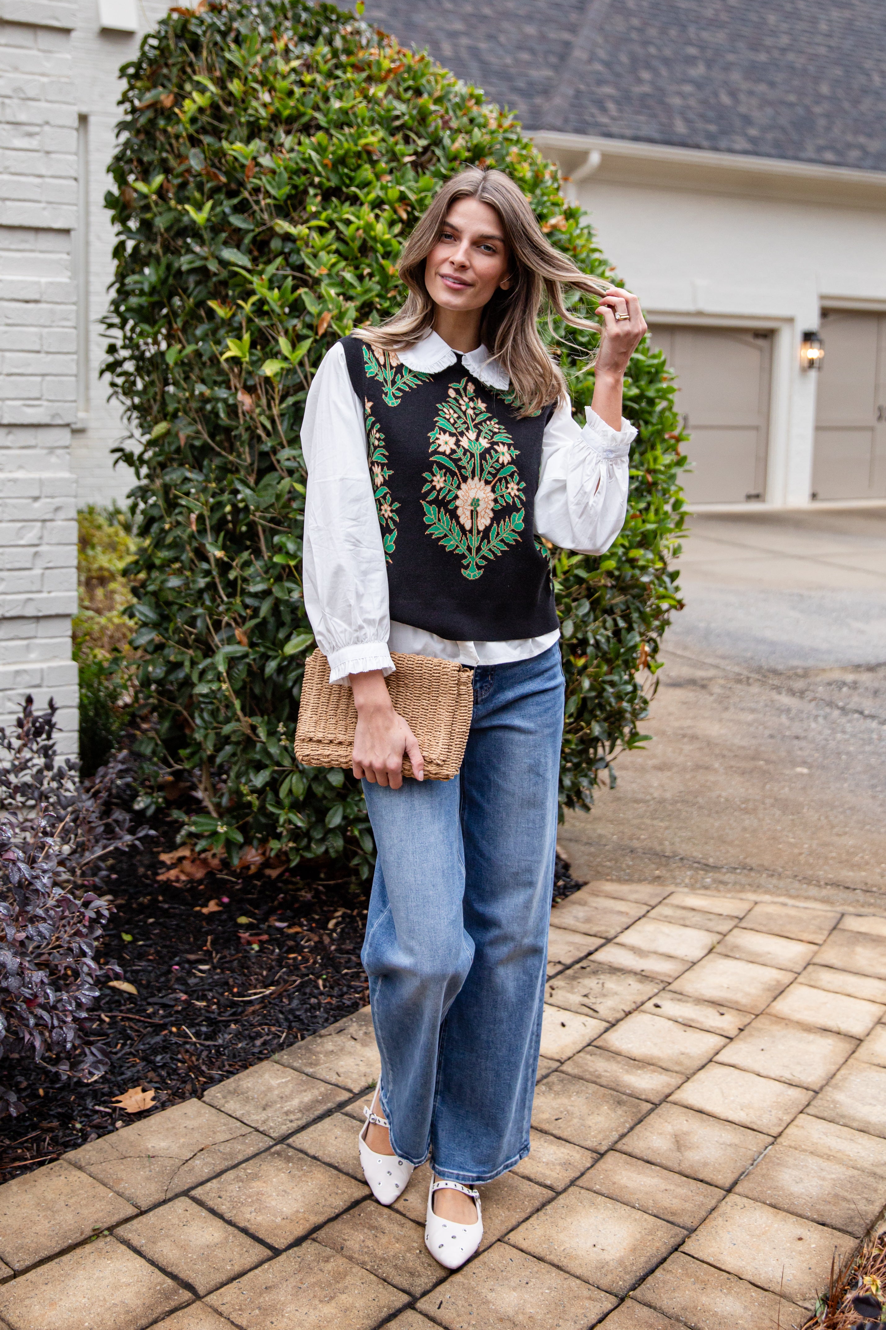 Woman in embroidered vest and blue jeans standing on a sidewalk with greenery and a house in the background.