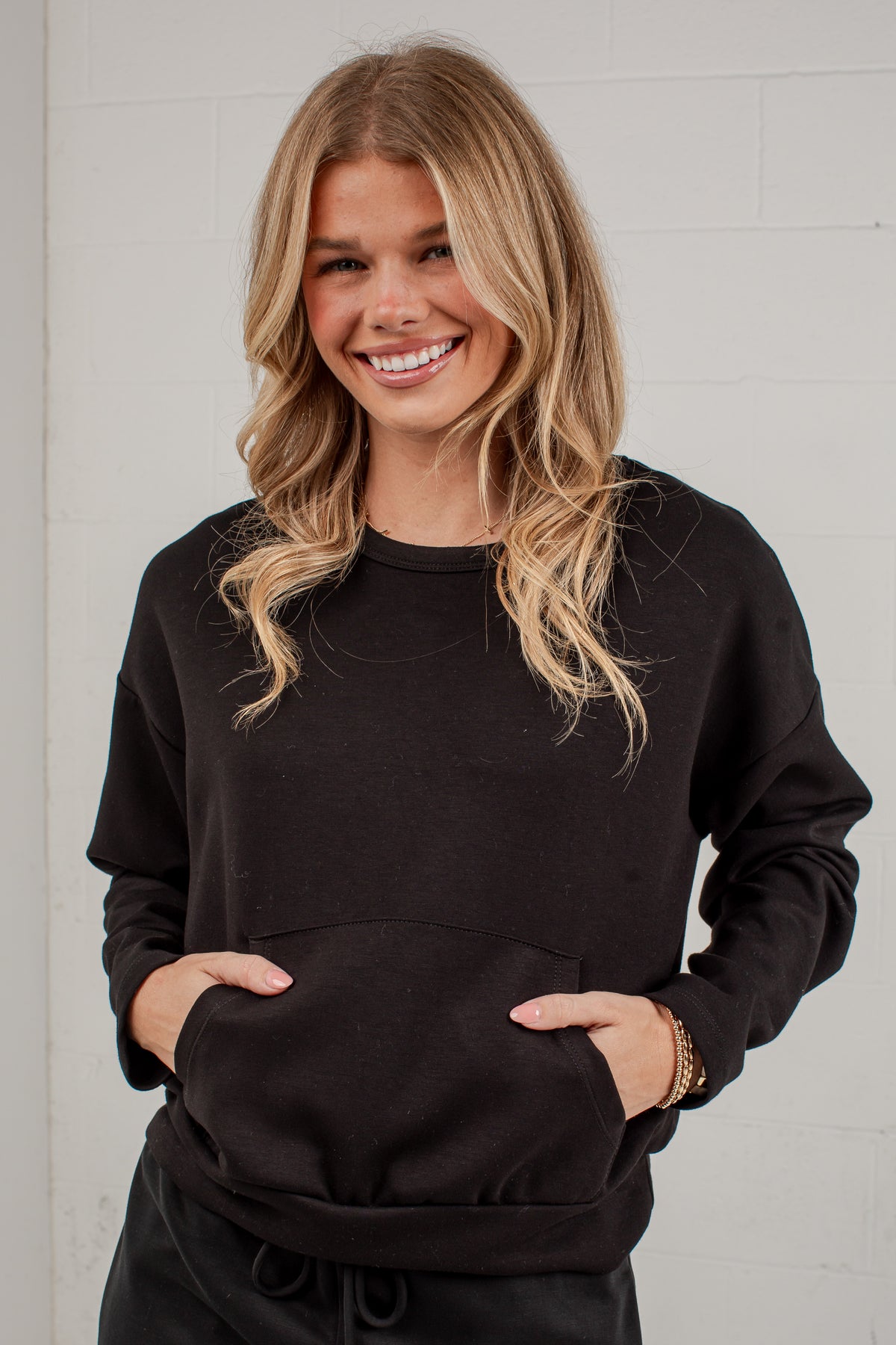 A smiling blonde woman wearing a black crewneck sweatshirt with a front kangaroo pocket, standing against a white brick wall.
