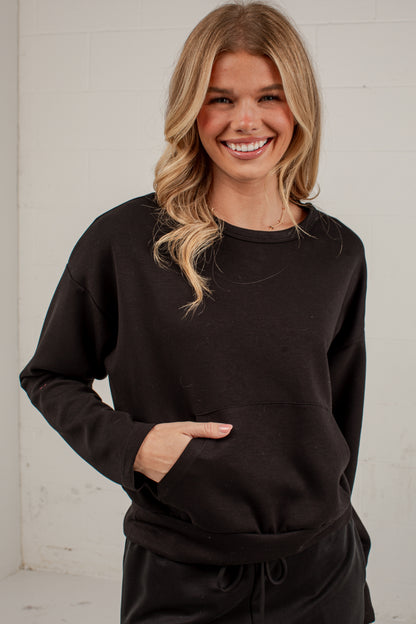 A smiling blonde woman wearing a black long-sleeve crewneck sweatshirt with a front kangaroo pocket, standing against a white brick wall.