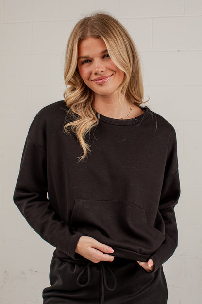 A woman with blonde hair wearing a black crewneck sweatshirt and matching black drawstring pants, standing against a white brick wall.