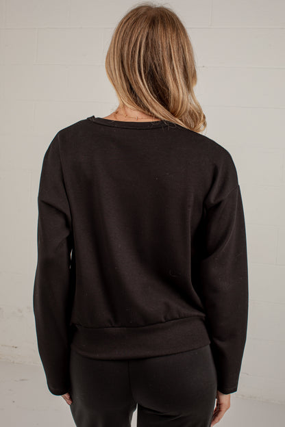 A woman viewed from behind wearing a dark brown crewneck sweatshirt and black pants against a white brick wall.