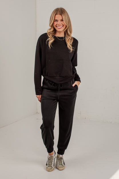 A woman modeling a black sweatshirt and jogger pants set with gray sneakers, standing against a white studio background.