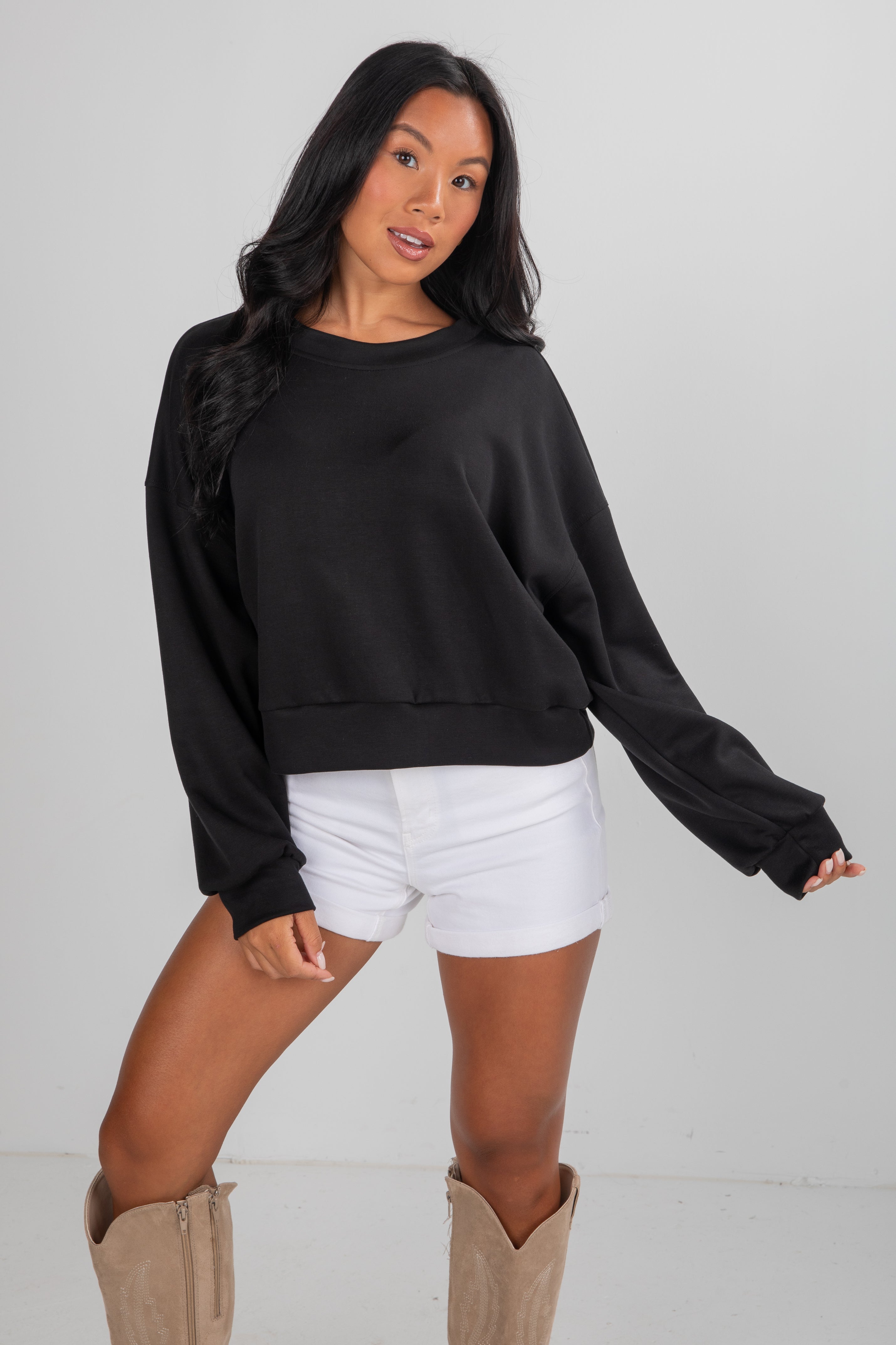 Woman wearing a black sweater and white shorts with beige boots on a plain background