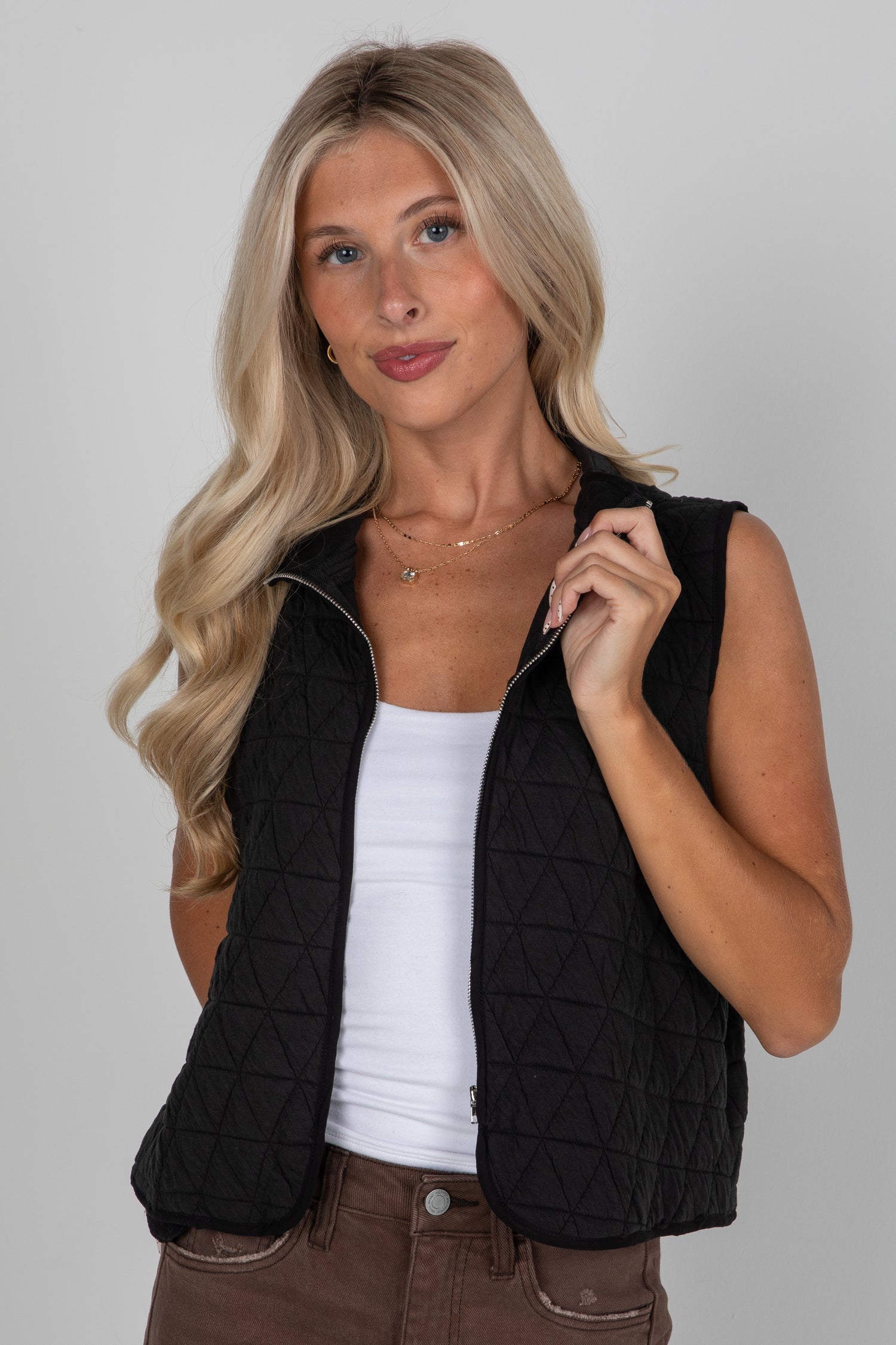 Woman wearing a black quilted vest over a white top and brown pants on a gray background