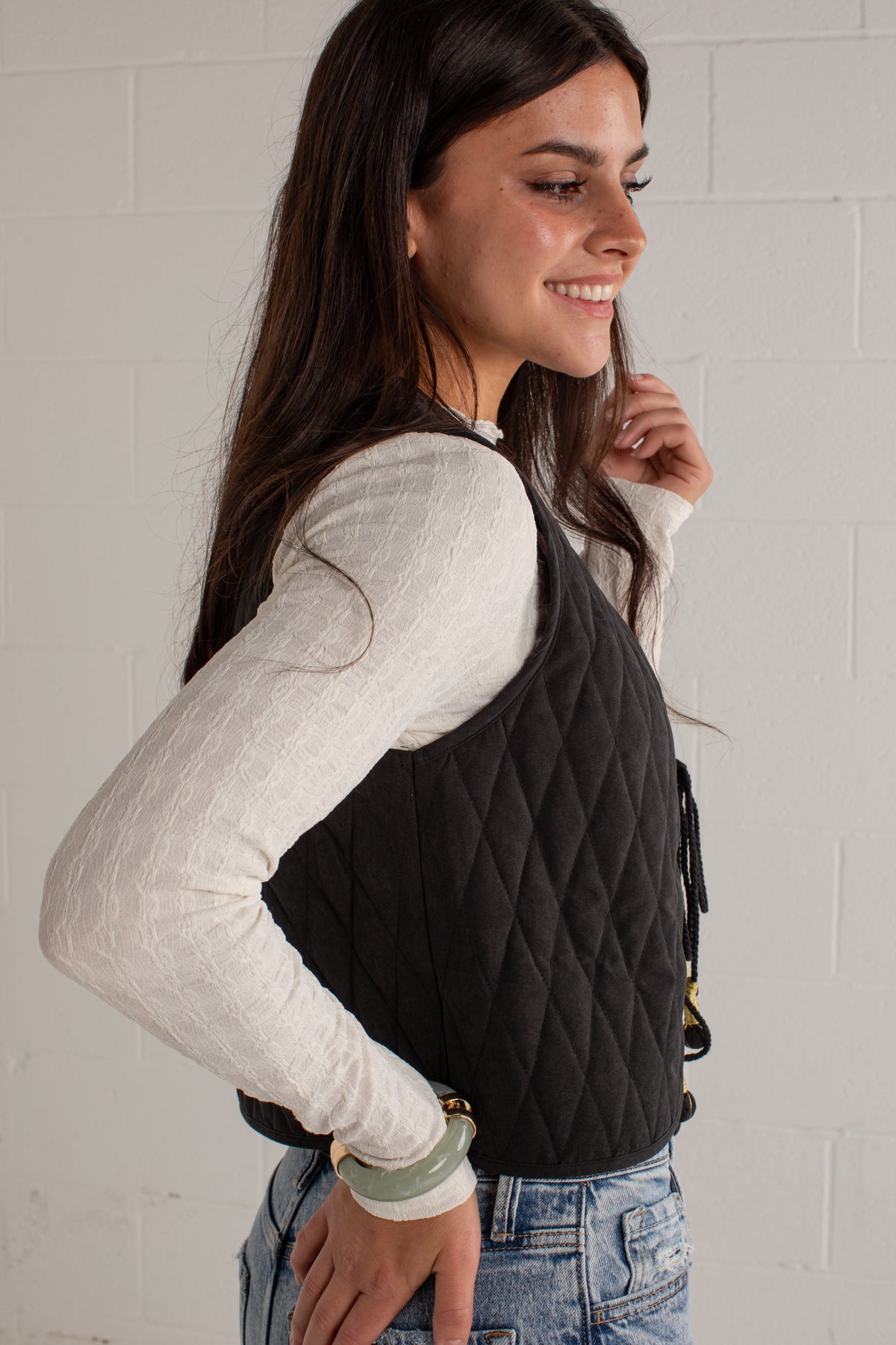 A woman wearing a black diamond-quilted sleeveless vest over a white textured long-sleeve top and blue jeans, standing against a white brick wall.