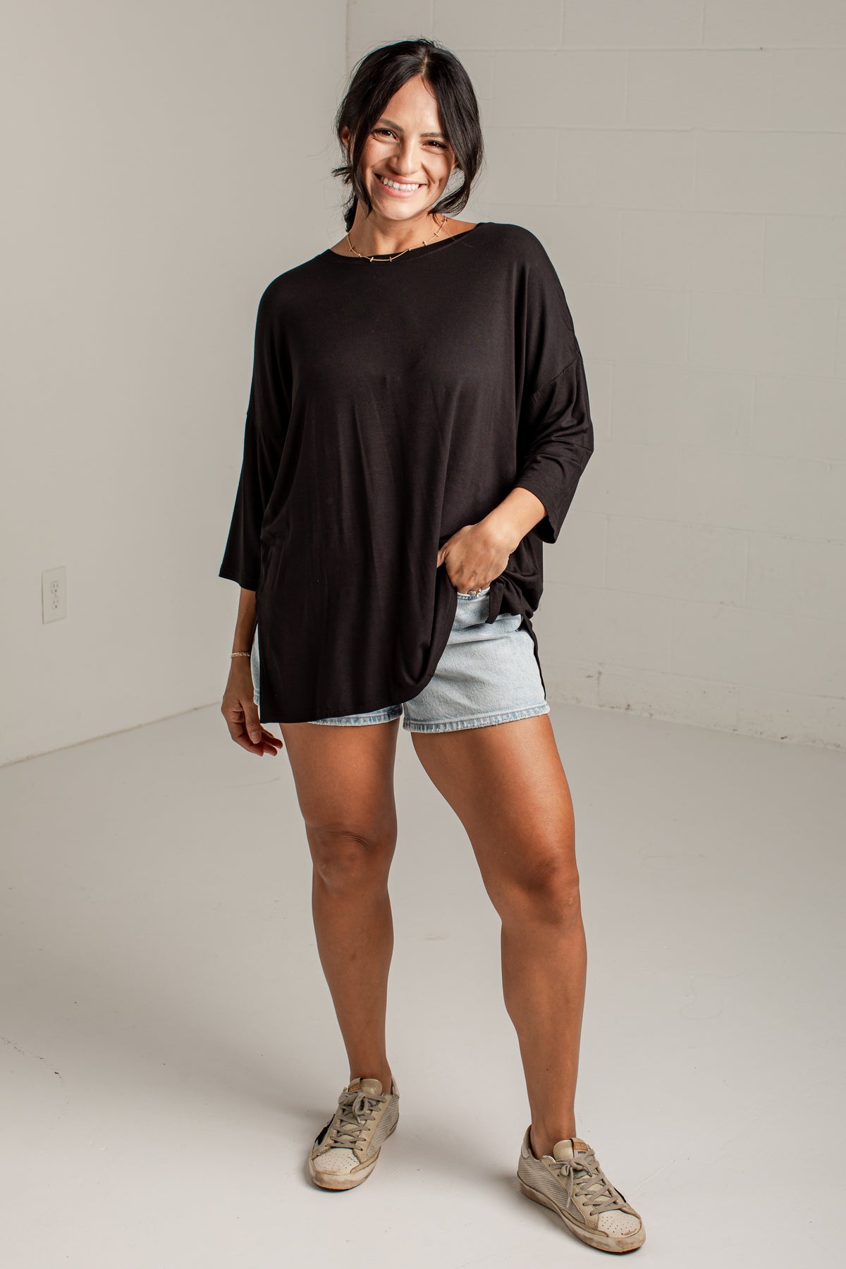 A woman modeling a loose-fitting black long-sleeve top with light denim shorts and beige sneakers, standing in a bright white studio.