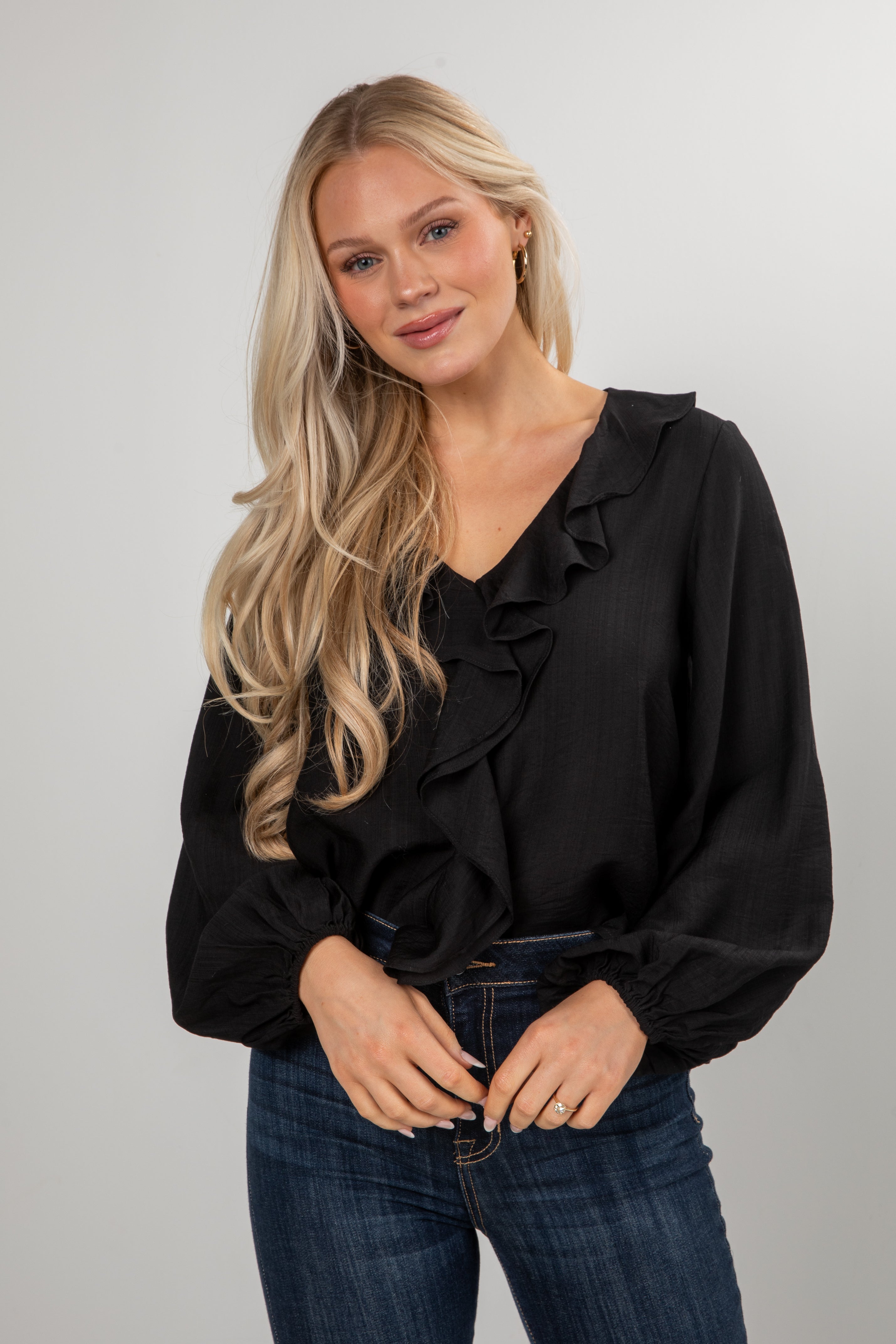 Woman wearing a black blouse with ruffles and blue jeans against a plain background