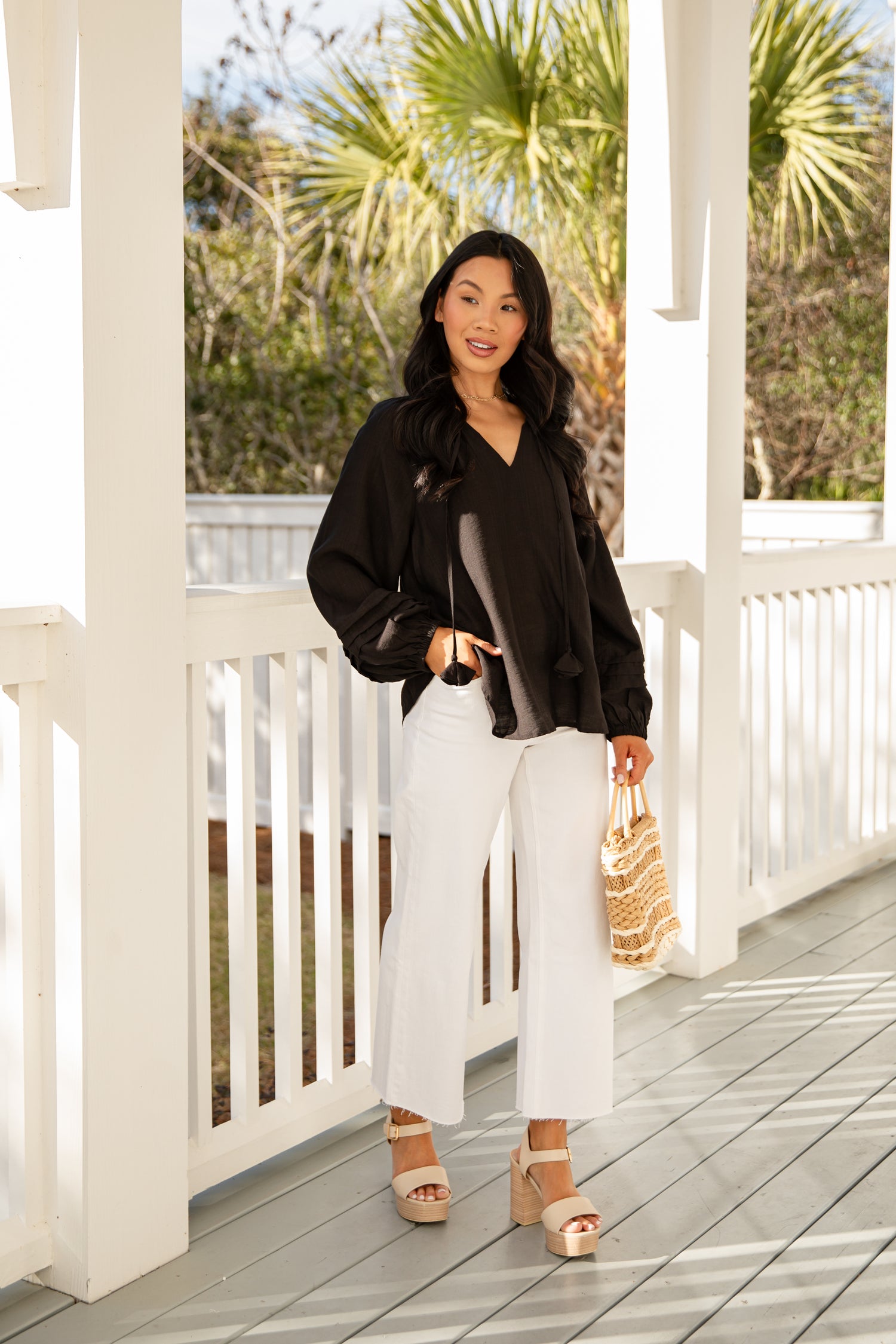Woman in black blouse and white pants standing on a wooden deck with palm trees in the background