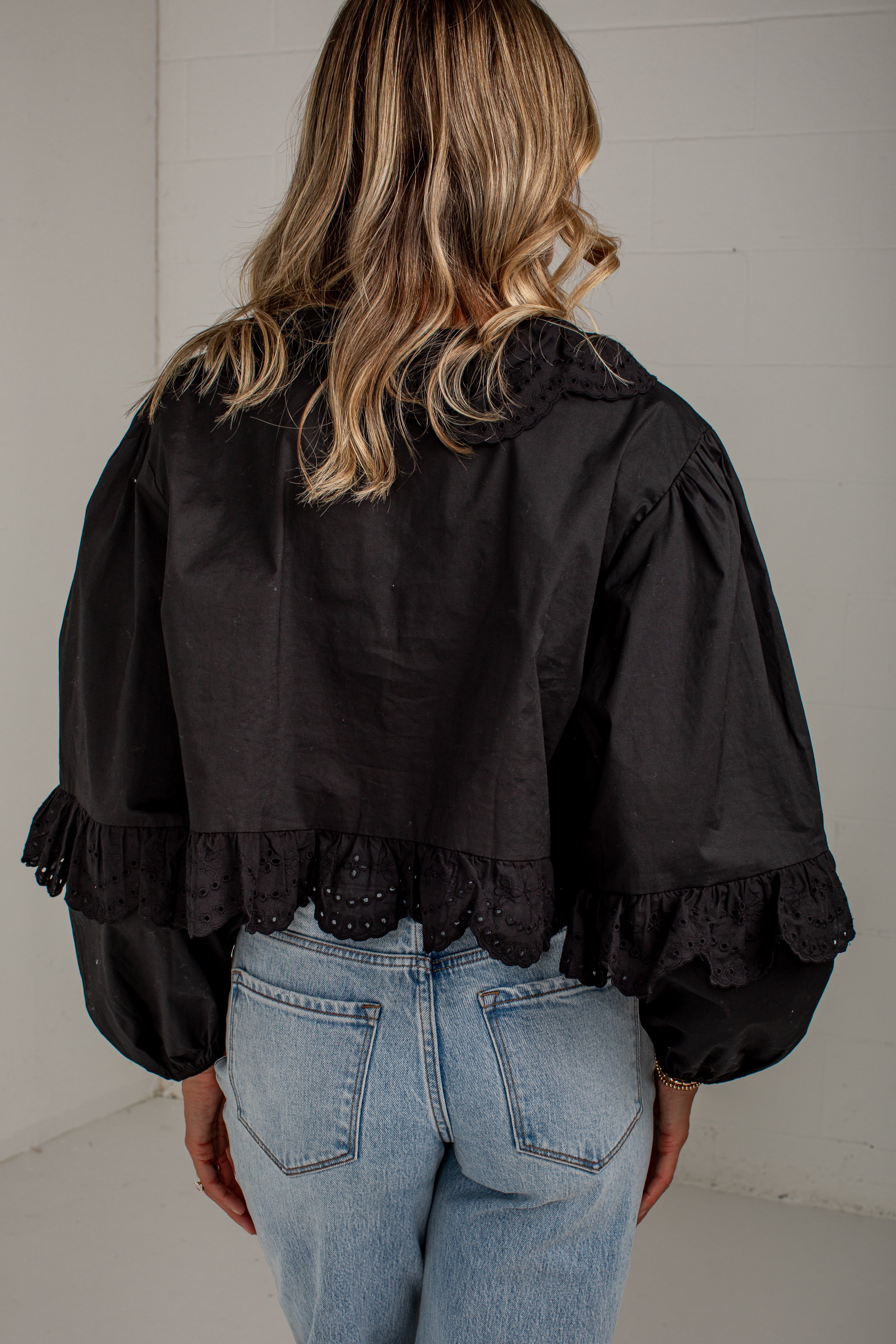 A woman viewed from behind wearing a black cropped blouse with dramatic puffed sleeves, lace trim at the collar and cuffs, and light blue jeans against a white paneled wall.