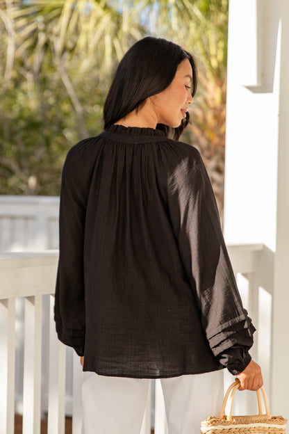 Woman wearing a black blouse with long sleeves and white pants, standing on a porch.