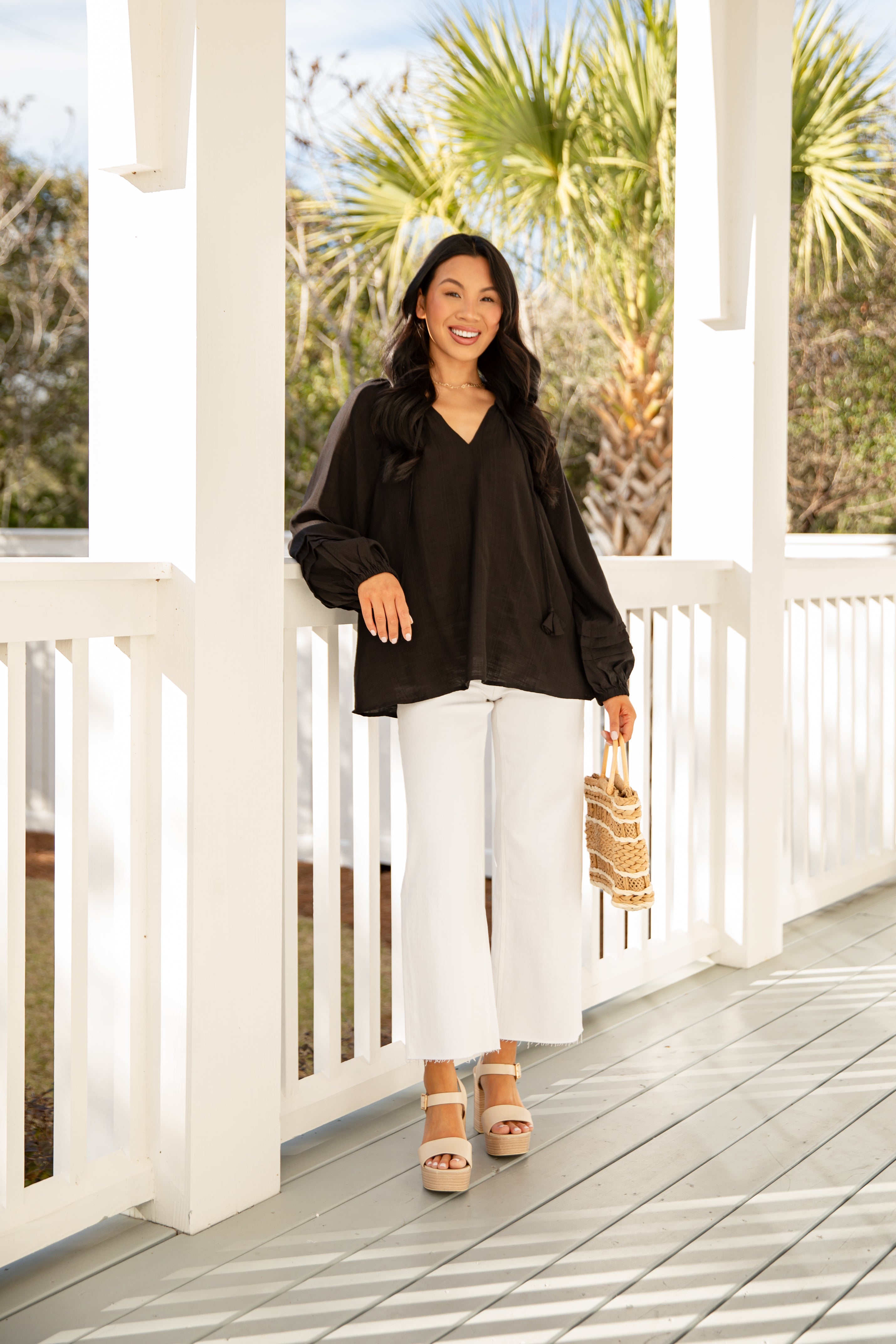 Woman in black top and white pants standing on a wooden deck with palm trees in the background