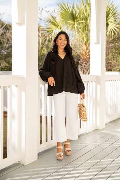Woman in black top and white pants standing on a wooden deck with palm trees in the background