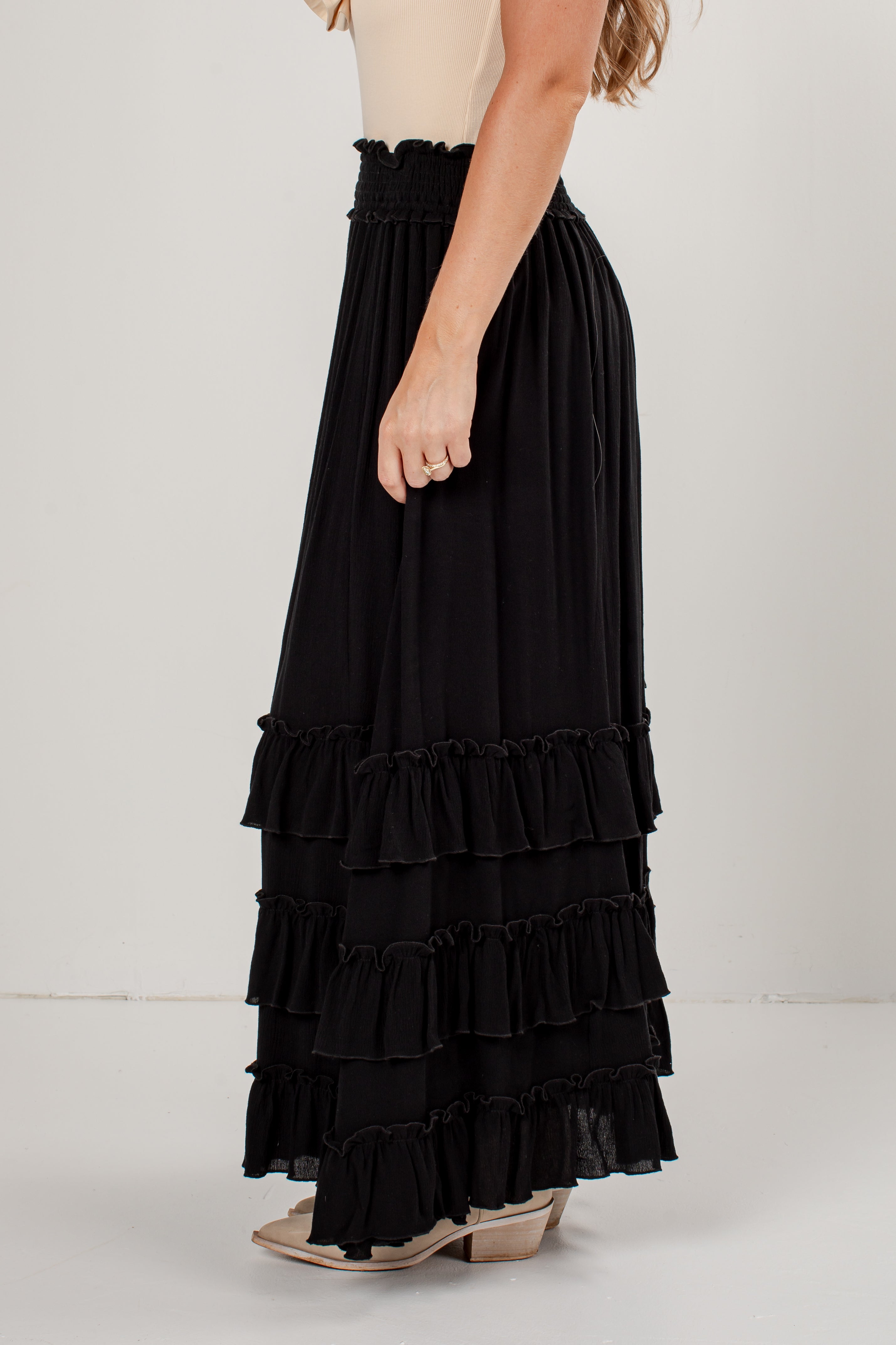 A black tiered maxi skirt with a smocked waistband, shown on a model from the side against a plain light gray studio background.