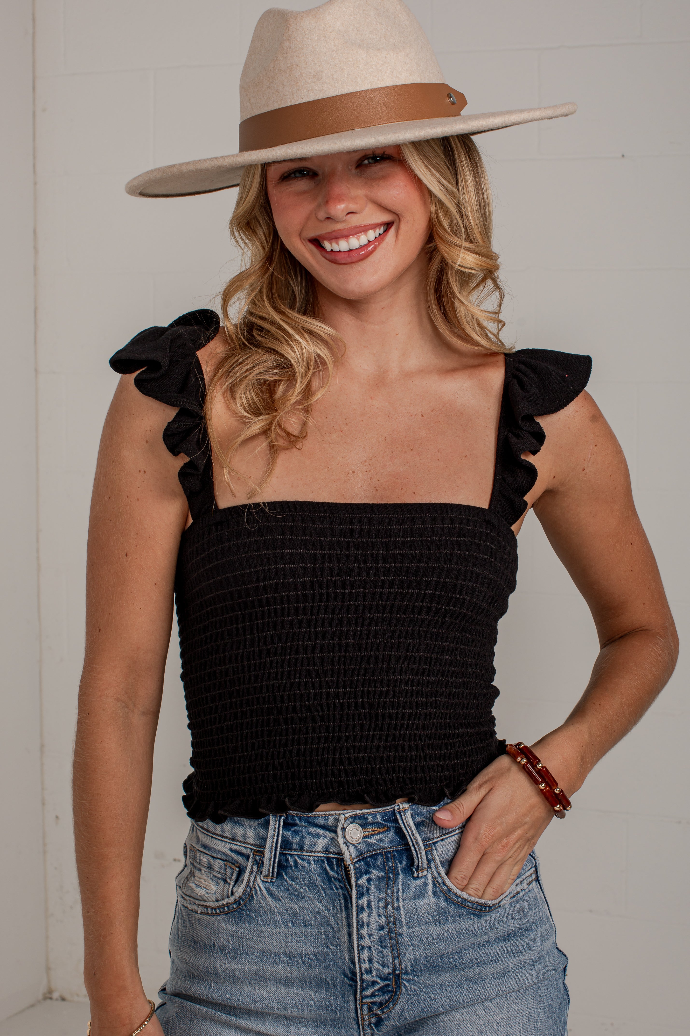 A smiling woman wearing a black smocked ruffle-strap top with blue jeans and a beige wide-brim hat, standing against a light gray wall.