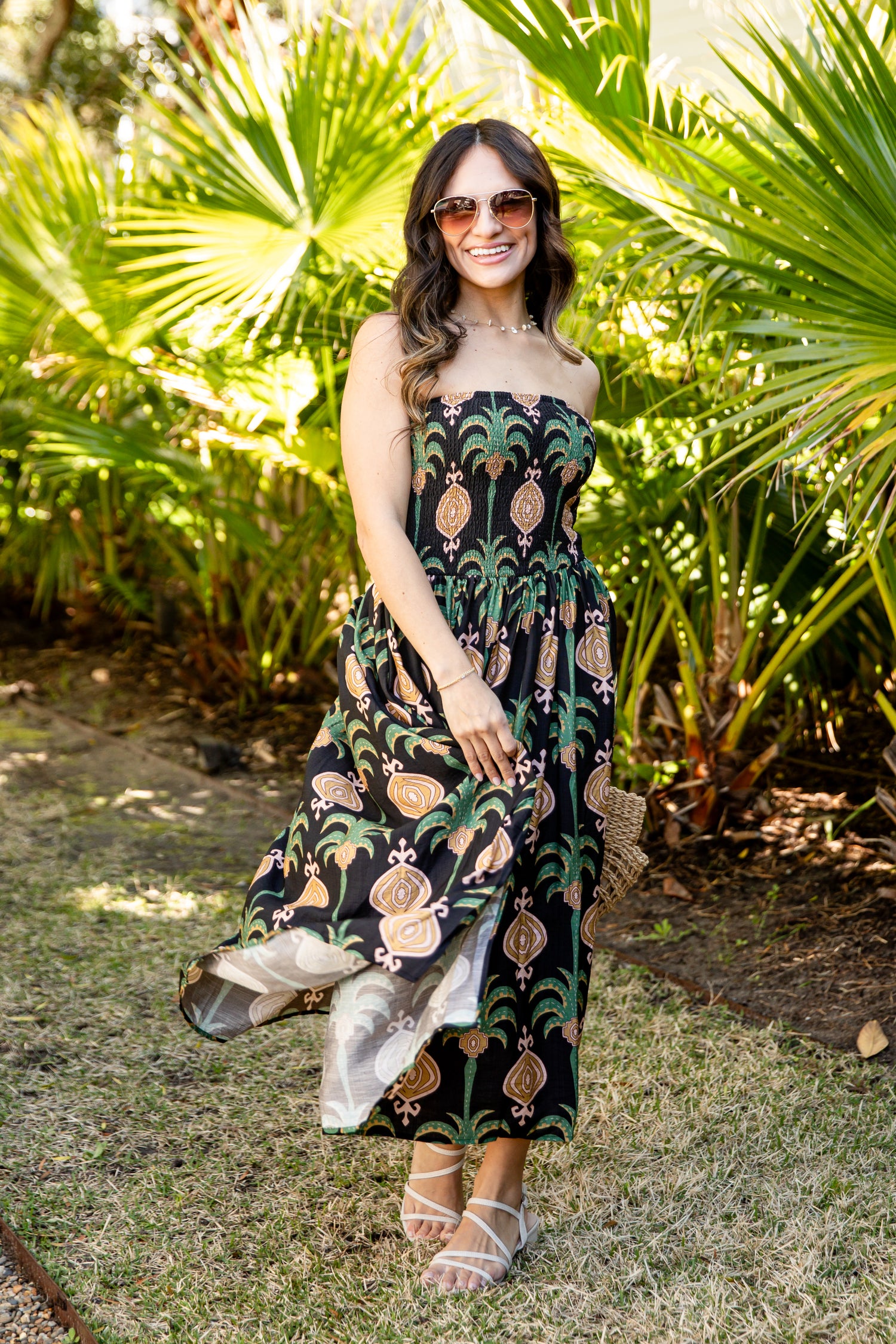 Woman in a strapless dress with a palm tree pattern standing in a garden with greenery.