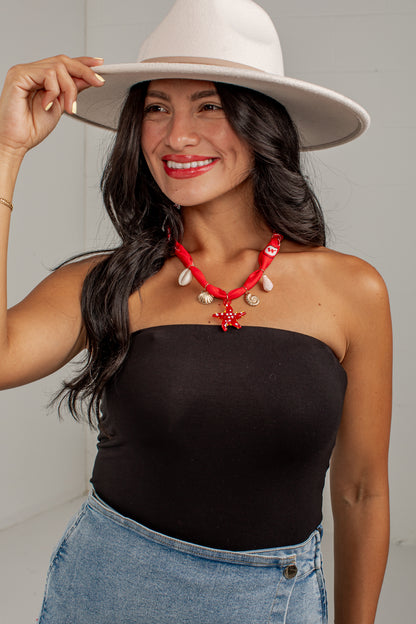 A woman wearing a black strapless tube top, a red and white shell necklace, a white wide-brim hat, and blue denim shorts, posing against a light gray wall.