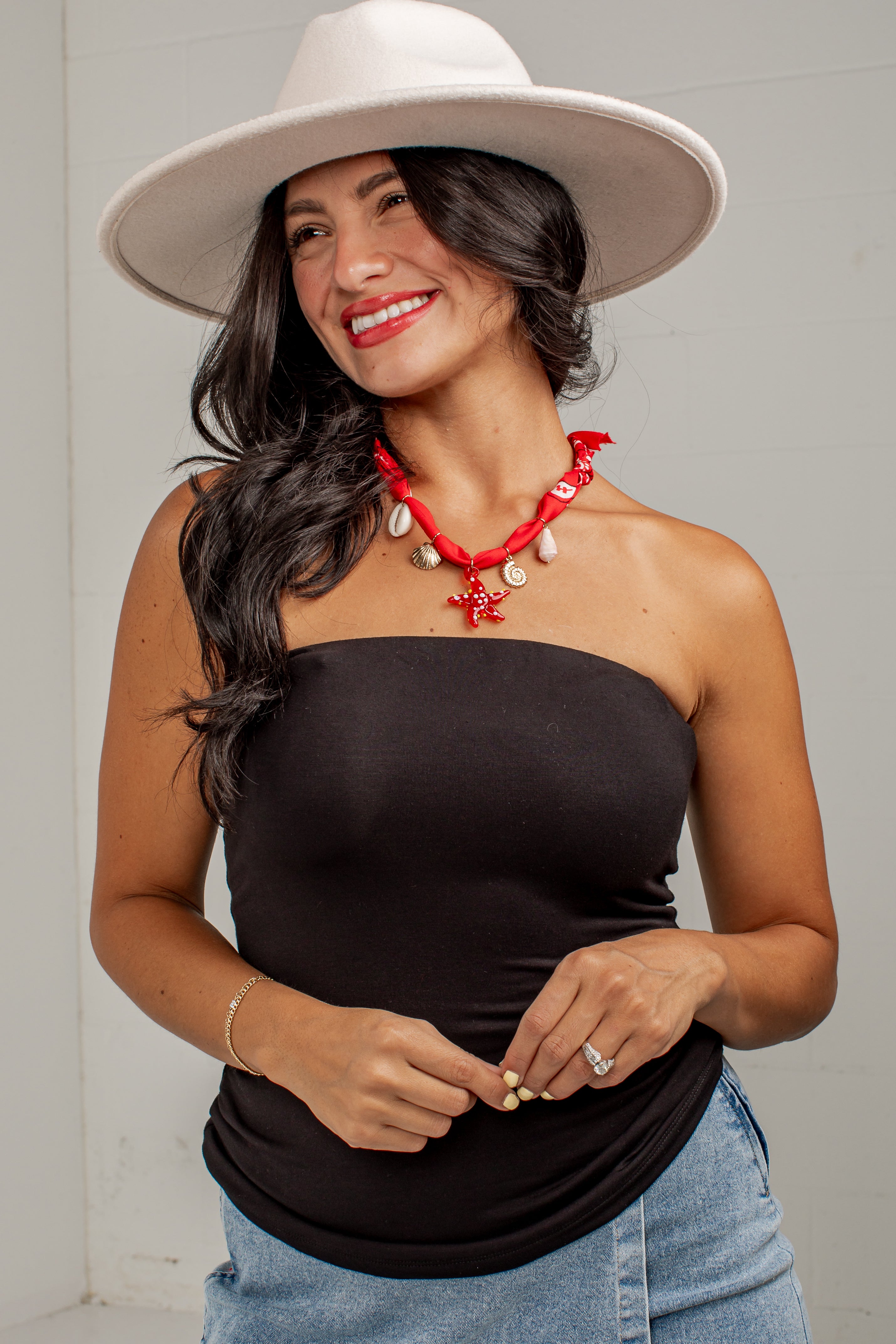 A woman wearing a black strapless tube top, blue jeans, a white wide-brim hat, and a red shell necklace, posing against a light gray wall.