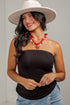 A woman wearing a black strapless tube top, blue jeans, a white wide-brim hat, and a red shell necklace, posing against a light gray wall.