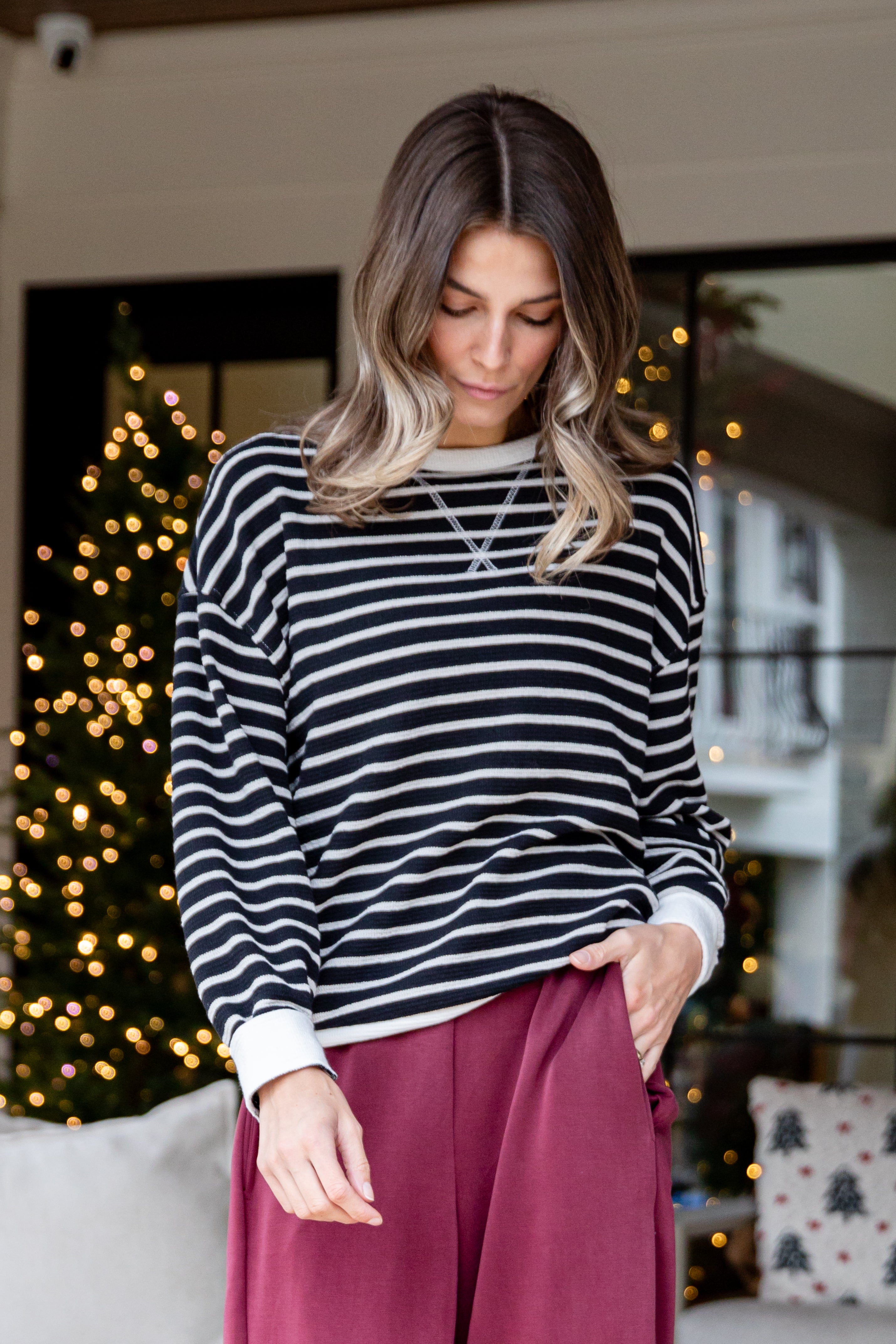 Woman wearing a striped sweater and burgundy pants in a festive indoor setting with Christmas lights.