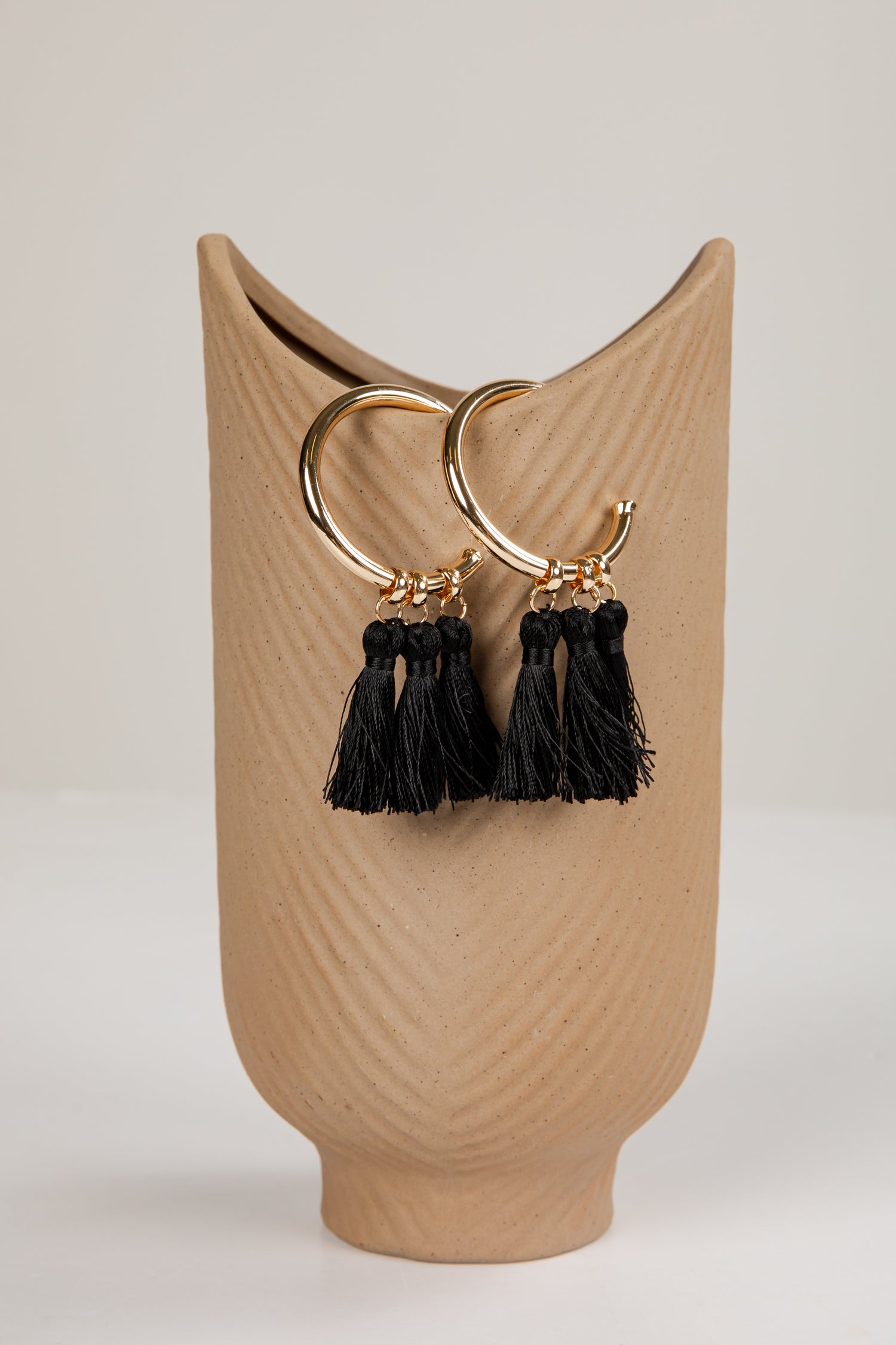 Gold hoop earrings with black tassels on a beige stand against a light background