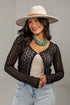 A woman wearing a black open-knit long-sleeve cardigan over a cream top, paired with light blue jeans and a cream wide-brim hat, against a plain light gray background.