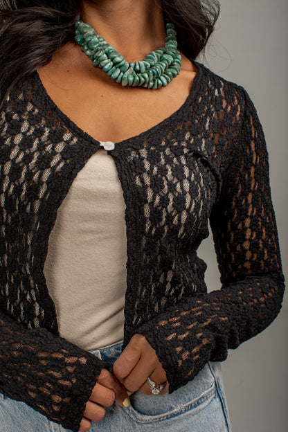 A close-up of a person wearing a black open-knit cardigan over a light beige top, paired with blue jeans and a chunky green beaded necklace, against a plain gray background.