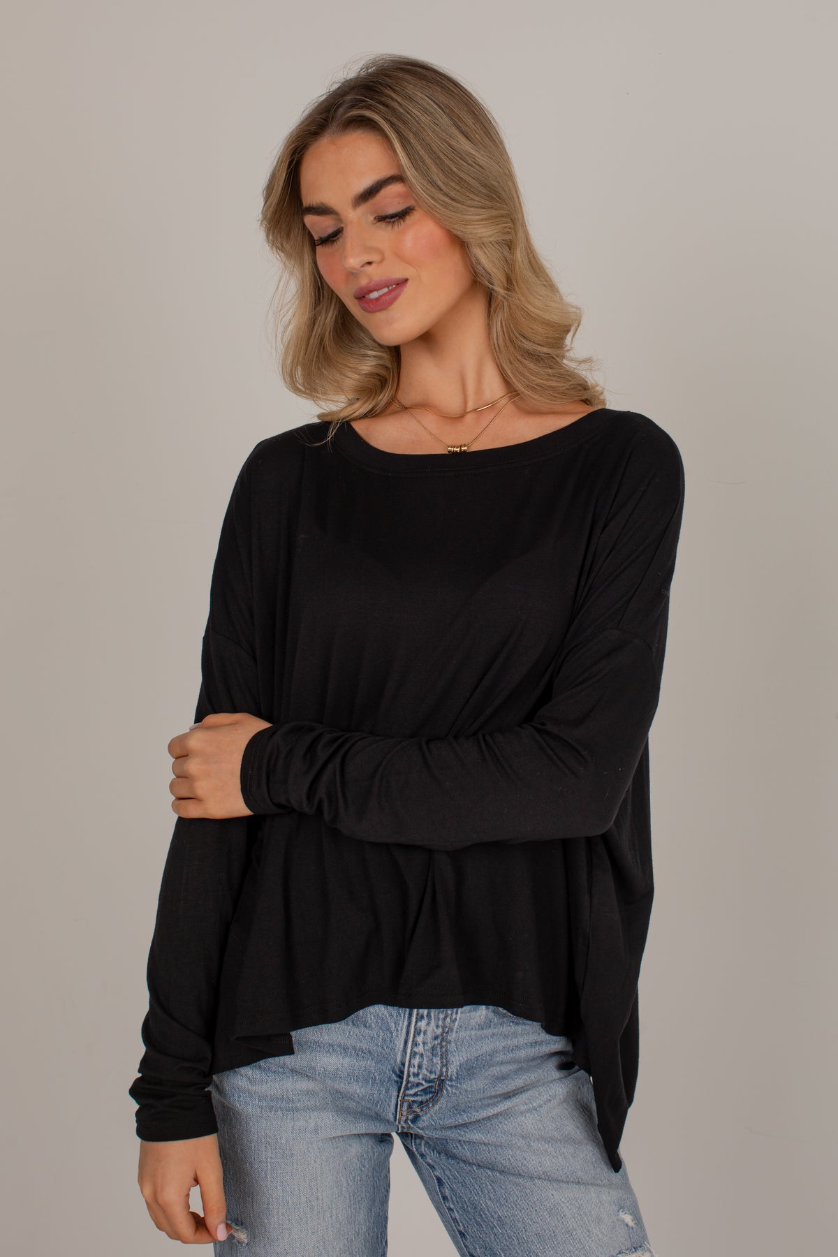 A woman wearing a black long-sleeve top with a wide boat neckline and a gathered peplum hem, paired with light-wash blue jeans, against a solid light gray studio background.