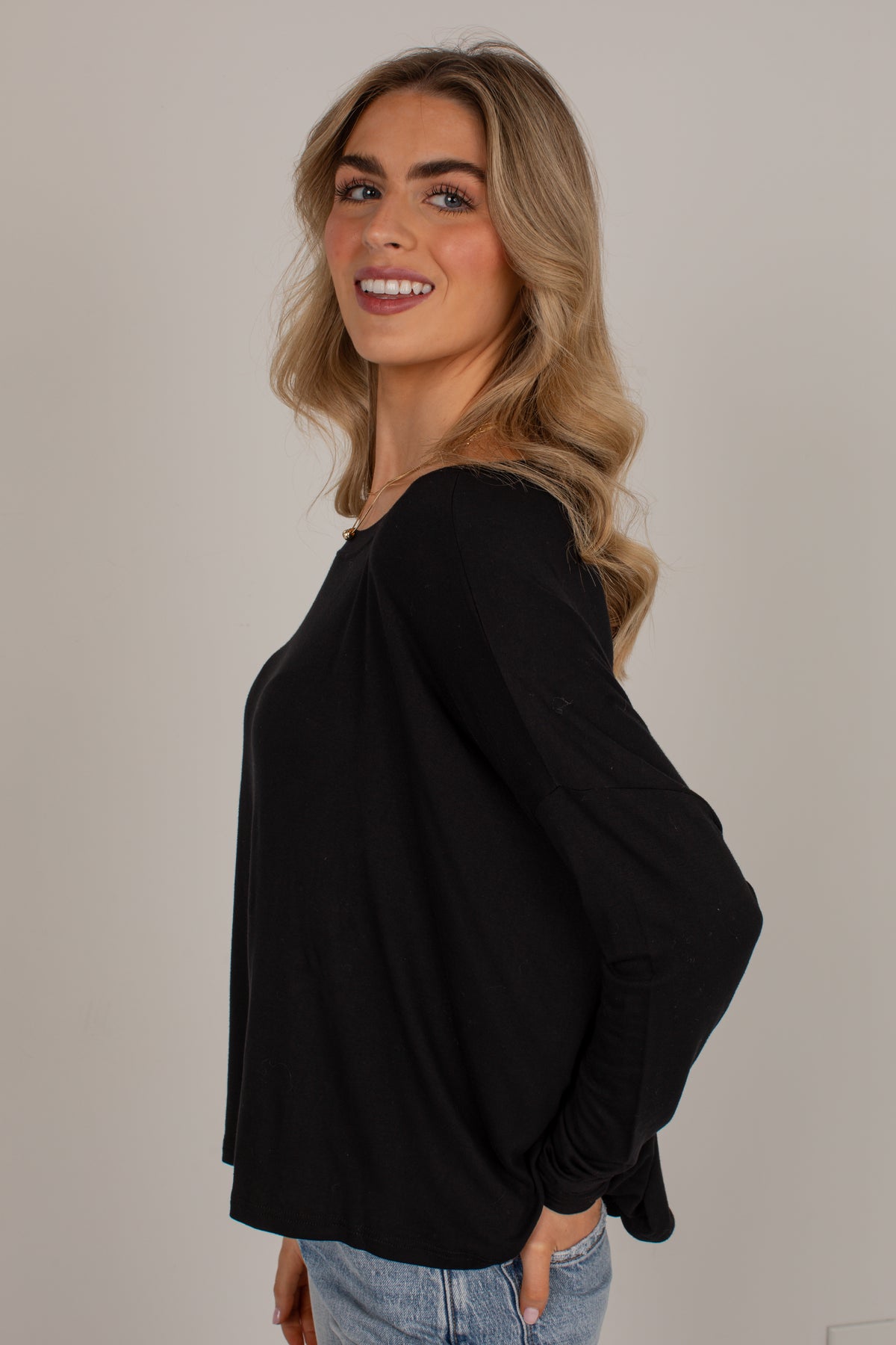 A woman with blonde hair wearing a black long-sleeve top and light blue jeans, posing against a neutral light gray background.
