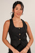 Woman wearing a black sleeveless top with gold buttons against a plain background