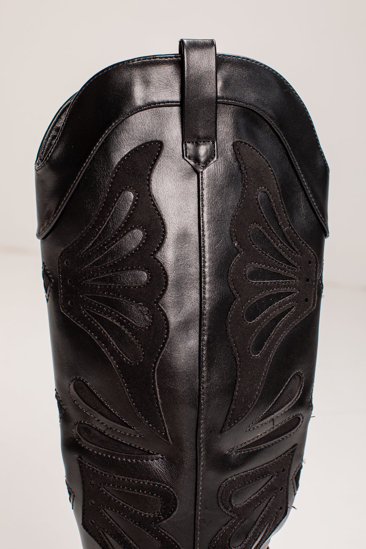 Close-up of a black leather cowboy boot shaft with decorative cutout stitching and a pull tab at the top, photographed against a plain light background.