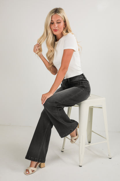 Woman sitting on a white stool wearing a white t-shirt and black jeans against a plain background