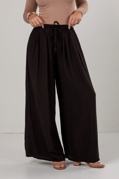 A person wearing loose, wide-leg black pants with an elastic waistband and drawstring, paired with a beige fitted top and heeled sandals, photographed against a plain light gray studio background.