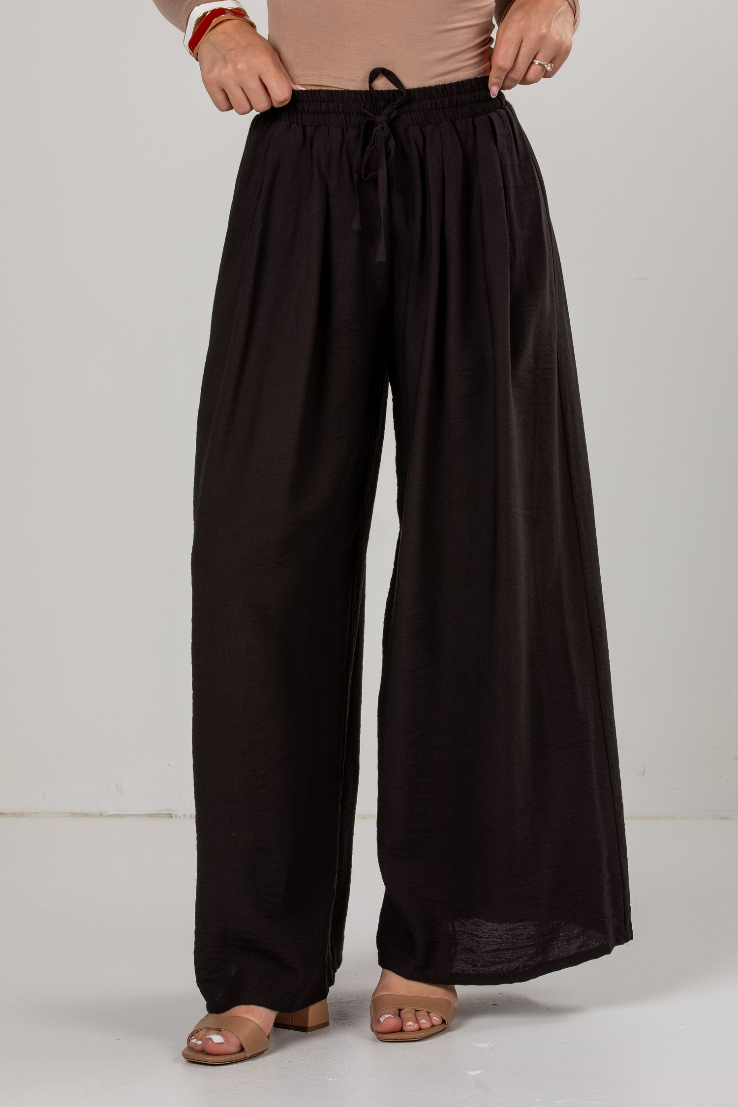 A person wearing loose, wide-leg black drawstring pants with a beige top and tan heeled sandals, standing against a plain light gray studio background.