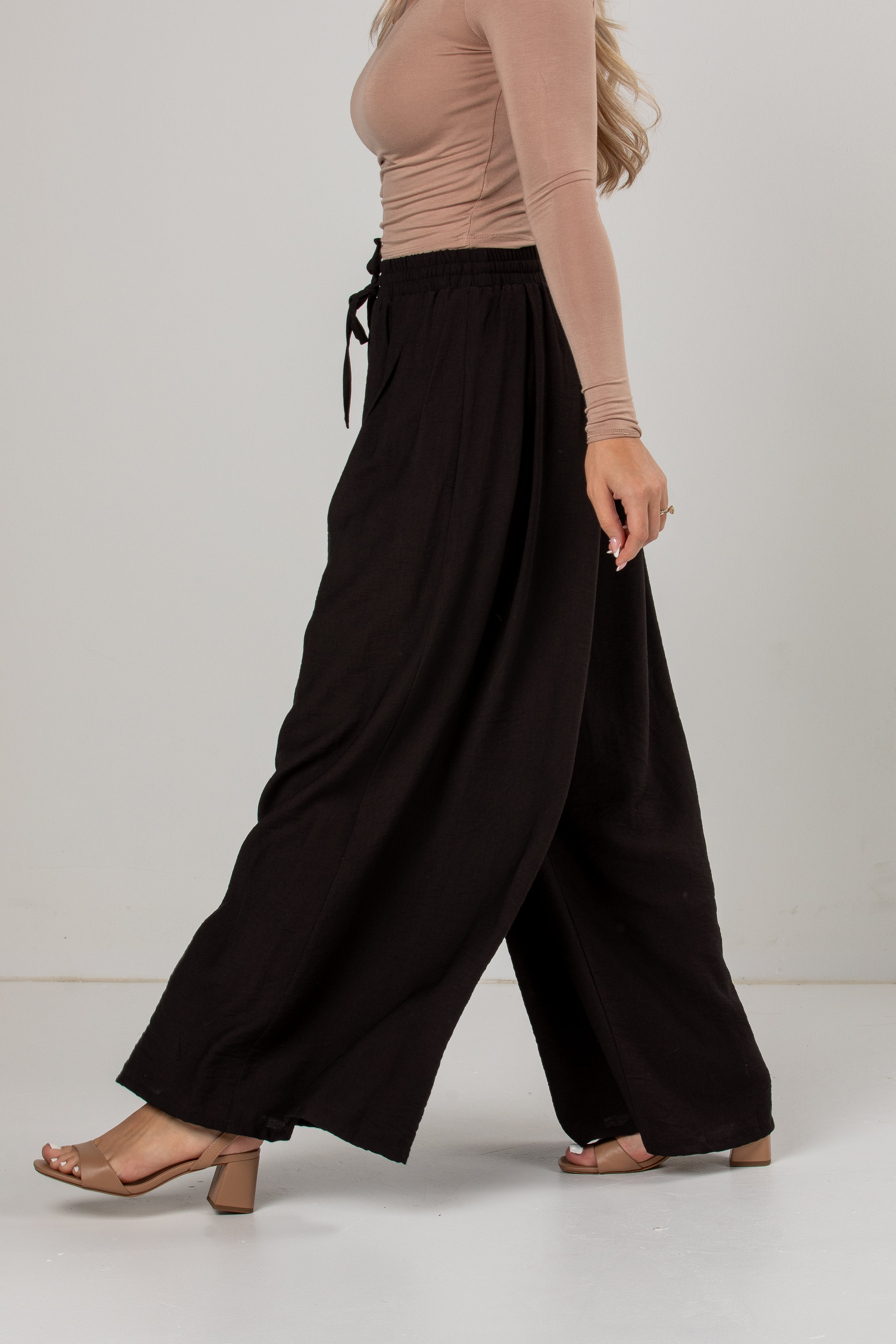 A model wearing black wide-leg drawstring pants, a beige long-sleeve top, and tan block-heel sandals, shown from the waist down in a side profile against a neutral studio background.