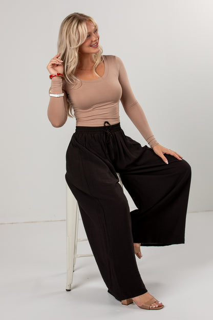 A woman models a beige long-sleeve scoop-neck top and black wide-leg drawstring pants, sitting on a white stool against a light gray studio background.